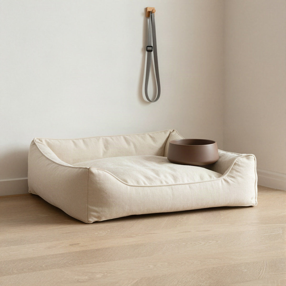 Cream dog bed with a brown ceramic bowl beside a hanging leash in a sunlit hallway